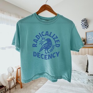 Peut inclure: Un t-shirt bleu sarcelle avec les mots "RADICALIZED BY BASIC DECENCY" en bleu. Un graphisme d'oiseau bleu avec une branche est au centre du texte. Le t-shirt est suspendu &agrave; un cintre en bois.