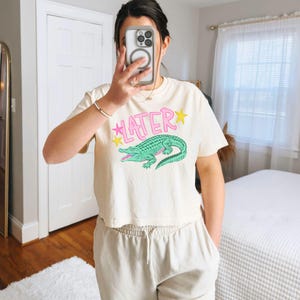 Peut inclure: Haut court cr&egrave;me avec le mot "LATER" en rose et un motif d'alligator vert. Le design comprend des &eacute;toiles jaunes. Le haut est port&eacute; avec un pantalon de surv&ecirc;tement beige clair.