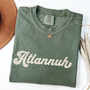 May include: An olive green t-shirt with the word "Atlannuh" in a retro font. A gold necklace with a pendant rests on the shirt. The shirt has a Comfort Colors tag.