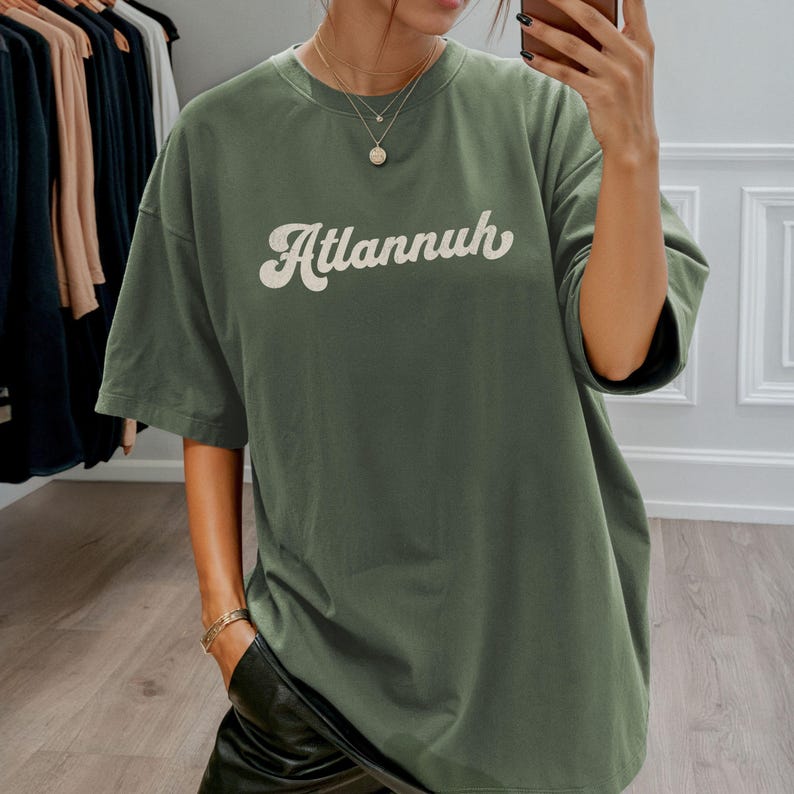 May include: An olive green t-shirt with the word "Atlannuh" in a white, retro-style font. The shirt has short sleeves and a relaxed fit. The person is wearing black pants and jewelry.