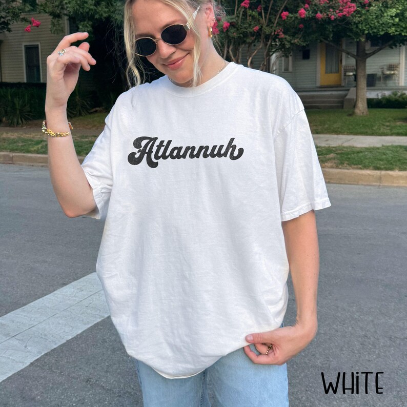 May include: White t-shirt with the word "Atlannuh" in a black, retro font. The shirt is a casual style, with short sleeves and a relaxed fit. The word "WHITE" is printed in the bottom right corner.