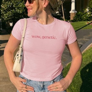 May include: A light pink short-sleeved t-shirt featuring the text "wow, genetic." printed in a dark red font across the chest. The shirt has a crew neckline and a fitted silhouette, paired with a white shoulder bag and blue denim jeans.