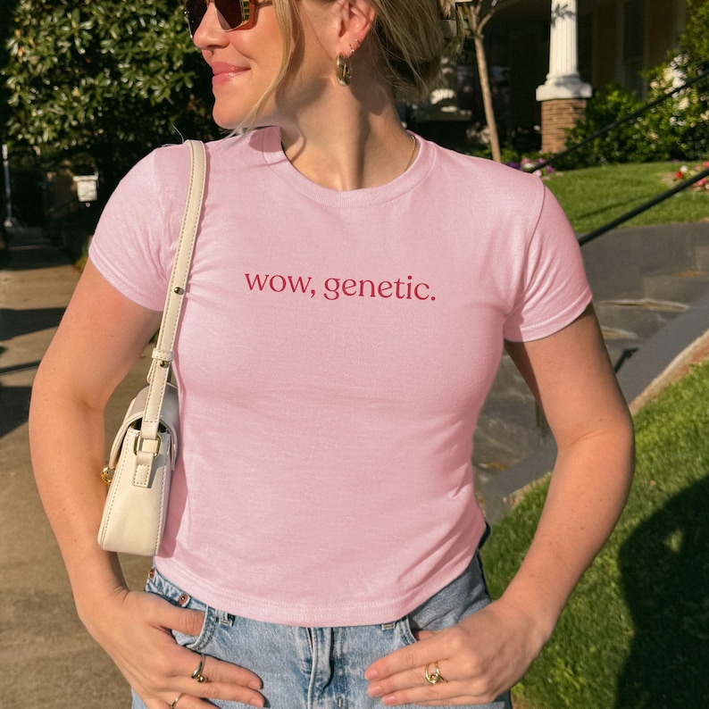 May include: A light pink short-sleeved t-shirt featuring the text "wow, genetic." printed in a dark red font across the chest. The shirt has a crew neckline and a fitted silhouette, paired with a white shoulder bag and blue denim jeans.