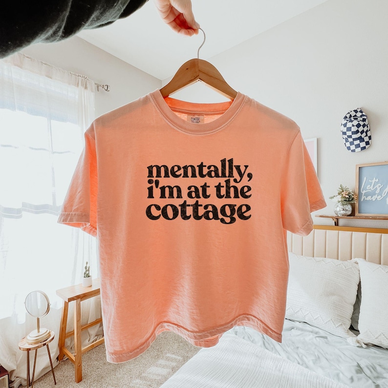 Op de afbeelding: Perzikkleurig T-shirt met de zwarte tekst "mentally, I'm at the cottage". Het shirt hangt aan een houten hanger. De achtergrond toont een bed, een kleine tafel en decoratieve items.