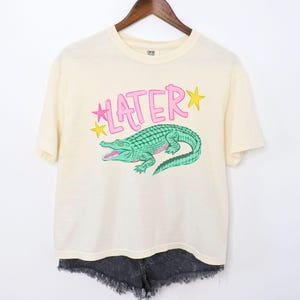 Peut inclure: T-shirt cr&egrave;me avec un motif d'alligator vert et le mot "LATER" en rose, avec des &eacute;toiles jaunes. Le t-shirt est suspendu &agrave; un cintre en bois, associ&eacute; &agrave; un short en jean noir.