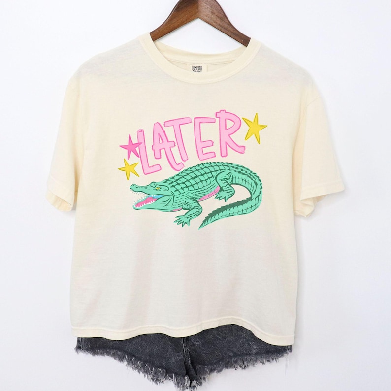 Peut inclure: T-shirt cr&egrave;me avec un motif d'alligator vert et le mot "LATER" en rose, avec des &eacute;toiles jaunes. Le t-shirt est suspendu &agrave; un cintre en bois, associ&eacute; &agrave; un short en jean noir.