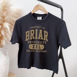 May include: A navy blue t-shirt with the text "PROPERTY OF BRIAR UNIVERSITY - XXL - ATHLETIC DEPARTMENT" in gold lettering. The shirt is hanging on a wooden hanger against a neutral background.