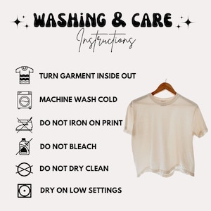 May include: A cream-colored t-shirt hanging on a wooden hanger, with washing instructions. The text reads: Turn garment inside out, machine wash cold, do not iron on print, do not bleach, do not dry clean, and dry on low settings.