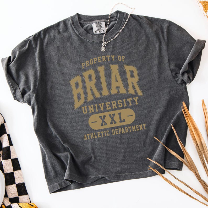 May include: A charcoal gray t-shirt with the text "PROPERTY OF BRIAR UNIVERSITY - XXL - ATHLETIC DEPARTMENT" in gold lettering. A silver necklace with a pendant is draped on the shirt.