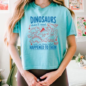 May include: A turquoise t-shirt with a playful dinosaur graphic. The design features various red dinosaur illustrations, books, and the text "DINOSAURS didn't read and we know what HAPPENED TO THEM" in blue. The shirt is a casual, comfortable style.
