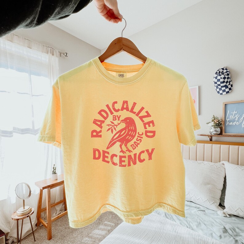Peut inclure: T-shirt jaune avec l'inscription "RADICALIZED BY BASIC DECENCY" dans un motif circulaire rouge repr&eacute;sentant un oiseau tenant une branche. Le t-shirt est suspendu &agrave; un cintre en bois.