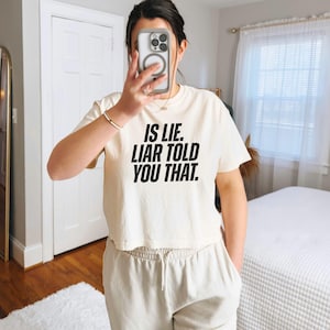 K&ouml;nnte beinhalten: Cremefarbenes, gecropptes T-Shirt mit dem Text "IS LIE. LIAR TOLD YOU THAT." in fetten schwarzen Buchstaben. Das Shirt wird mit hellbeigen Jogginghosen kombiniert. Die Person h&auml;lt ein Telefon vor ihr Gesicht.
