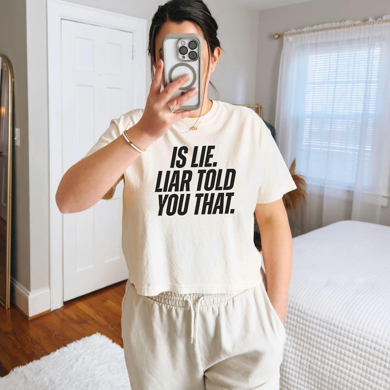 K&ouml;nnte beinhalten: Cremefarbenes, gecropptes T-Shirt mit dem Text "IS LIE. LIAR TOLD YOU THAT." in fetten schwarzen Buchstaben. Das Shirt wird mit hellbeigen Jogginghosen kombiniert. Die Person h&auml;lt ein Telefon vor ihr Gesicht.