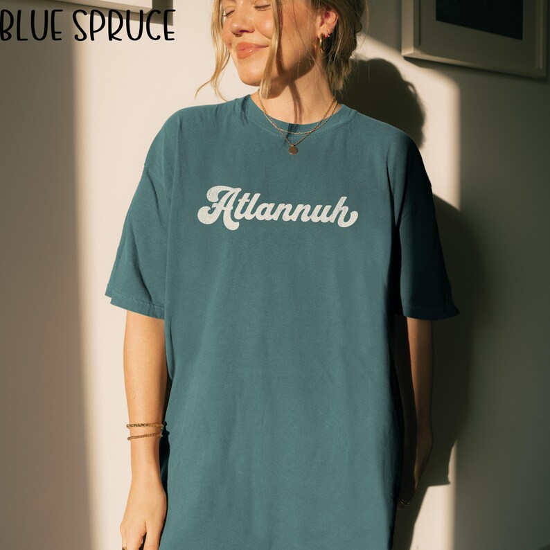 May include: Teal-colored t-shirt with the word "Atlannuh" in a white, retro font. The shirt has short sleeves and a relaxed fit. The brand name "Blue Spruce" is in the top left corner.