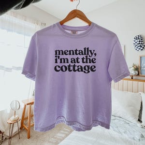 Op de afbeelding: Lichtpaars T-shirt met de zwarte tekst "mentally, I'm at the cottage". Het shirt hangt aan een houten hanger.