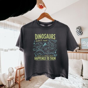 May include: Dark gray t-shirt with the text "DINOSAURS didn't read and we know what HAPPENED TO THEM" in green and blue. The shirt features dinosaur illustrations and book graphics. The shirt is hanging on a wooden hanger.