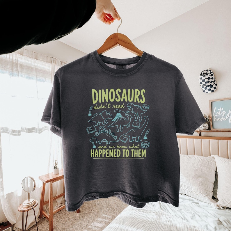 May include: Dark gray t-shirt with the text "DINOSAURS didn't read and we know what HAPPENED TO THEM" in green and blue. The shirt features dinosaur illustrations and book graphics. The shirt is hanging on a wooden hanger.