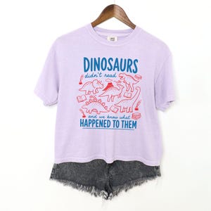 May include: A light purple t-shirt with the text "DINOSAURS didn't read and we know what HAPPENED TO THEM" in blue and red. The shirt features red illustrations of dinosaurs and books. The shirt is hanging on a wooden hanger.