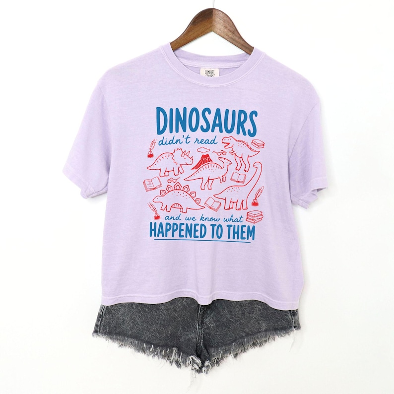 May include: A light purple t-shirt with the text "DINOSAURS didn't read and we know what HAPPENED TO THEM" in blue and red. The shirt features red illustrations of dinosaurs and books. The shirt is hanging on a wooden hanger.