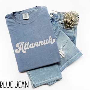 May include: A light blue t-shirt with the word "Atlannuh" in white script, paired with light blue denim jeans. The jeans have a distressed design and a small bouquet of white flowers in the pocket.