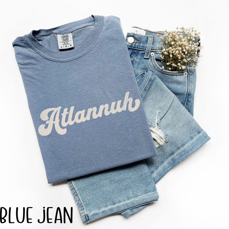 May include: A light blue t-shirt with the word "Atlannuh" in white script, paired with light blue denim jeans. The jeans have a distressed design and a small bouquet of white flowers in the pocket.