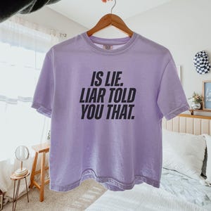 K&ouml;nnte beinhalten: Ein helllila T-Shirt mit dem Text "IS LIE. LIAR TOLD YOU THAT" in fetten schwarzen Buchstaben. Das Shirt h&auml;ngt an einem Holzb&uuml;gel. Der Hintergrund ist weich und neutral.