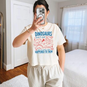 May include: Cream-colored, cropped t-shirt with the text "DINOSAURS didn't read, and we know what HAPPENED TO THEM" in blue and red. The shirt features dinosaur illustrations and books.
