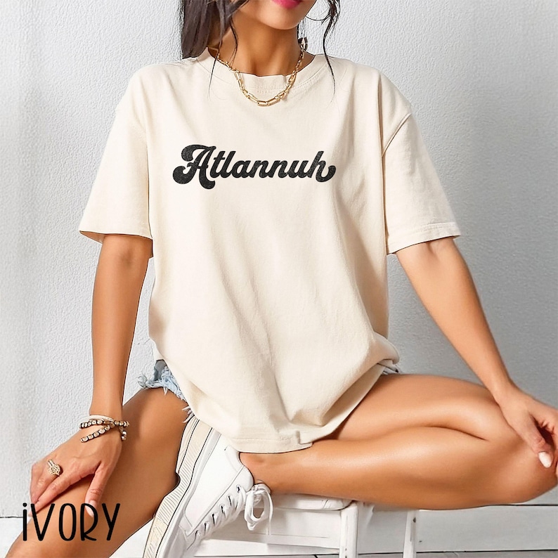 May include: Ivory-colored t-shirt with the word "Atlannuh" in a black, cursive font. The shirt has short sleeves and a relaxed fit. The model is wearing denim shorts and white sneakers.