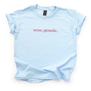 May include: A light blue short-sleeve t-shirt with a crew neckline and rolled sleeves. The center of the chest features the text "wow, genetic." printed in a dark red, lowercase serif font. The garment has a standard fit and a smooth texture.