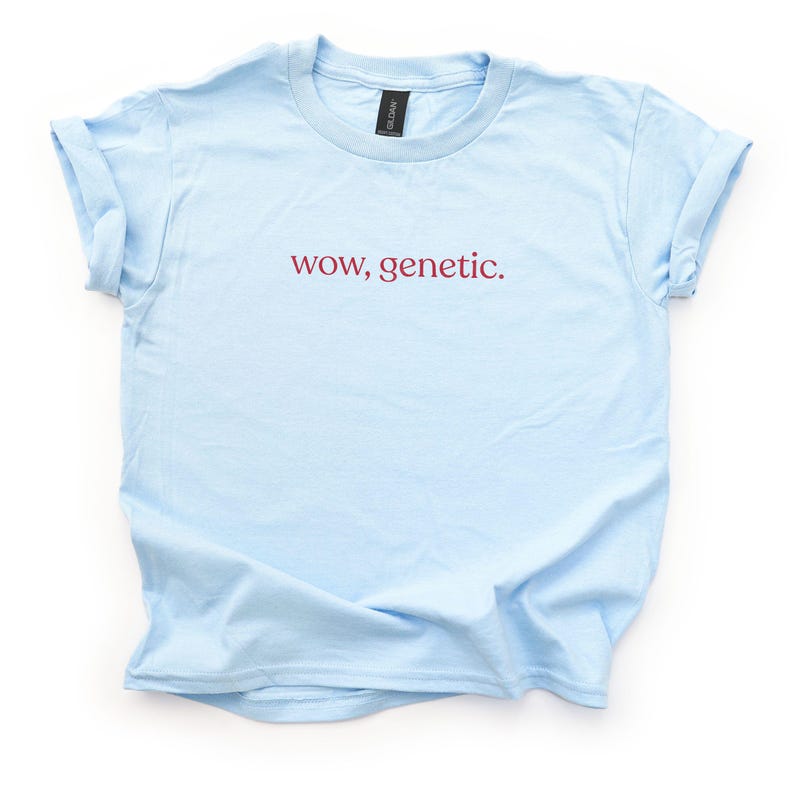 May include: A light blue short-sleeve t-shirt with a crew neckline and rolled sleeves. The center of the chest features the text "wow, genetic." printed in a dark red, lowercase serif font. The garment has a standard fit and a smooth texture.