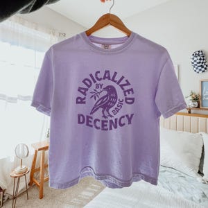 Peut inclure: T-shirt lilas avec l'inscription "RADICALIZED BY BASIC DECENCY" entourant un dessin de corbeau. Le t-shirt est suspendu &agrave; un cintre en bois.