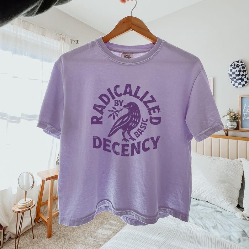 Peut inclure: T-shirt lilas avec l'inscription "RADICALIZED BY BASIC DECENCY" entourant un dessin de corbeau. Le t-shirt est suspendu &agrave; un cintre en bois.