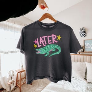 Peut inclure: T-shirt gris fonc&eacute; avec un motif d'alligator vert et le mot "LATER" en rose avec des &eacute;toiles jaunes. Le t-shirt est suspendu &agrave; un cintre en bois.
