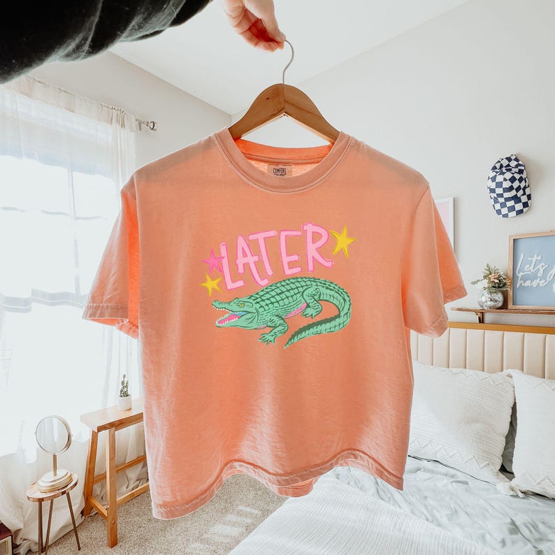 Peut inclure: T-shirt p&ecirc;che avec un motif d'alligator vert et le mot "LATER" en rose et jaune. Le t-shirt est suspendu &agrave; un cintre en bois.