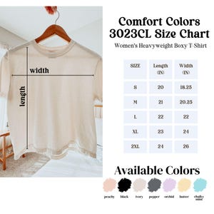 May include: A beige Comfort Colors 3023CL heavyweight boxy T-shirt hanging on a wooden hanger. The image includes a size chart with measurements in inches and a color palette featuring peachy, black, ivory, pepper, orchid, butter, and chalky mint.