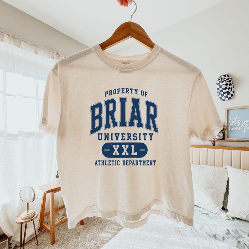 May include: Cream-colored t-shirt with the text "PROPERTY OF BRIAR UNIVERSITY -XXL- ATHLETIC DEPARTMENT" in navy blue. The shirt is hanging on a wooden hanger. The background includes a bed, a small table, and a window with a sheer curtain.