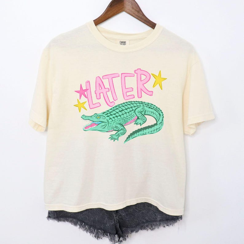 Puede incluir: Camiseta color crema con un gr&aacute;fico de un caim&aacute;n verde y la palabra "LATER" en rosa, con acentos de estrellas amarillas. La camiseta est&aacute; colgada en una percha de madera, combinada con shorts vaqueros negros.