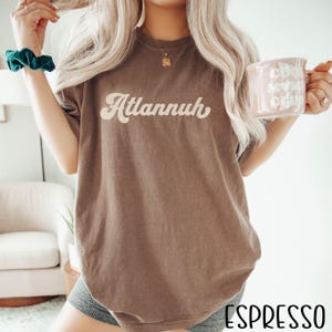 May include: Espresso-colored t-shirt with the word "Atlannuh" in a retro font. The shirt is a solid color and has a relaxed fit. The word "Espresso" is printed at the bottom of the image.