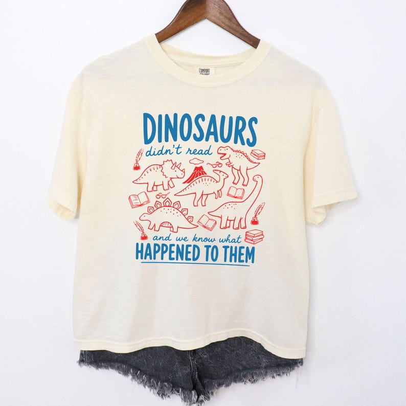 May include: Cream-colored t-shirt with a blue and red graphic. The graphic features dinosaur illustrations and the text "DINOSAURS didn't read and we know what HAPPENED TO THEM". The shirt is hanging on a wooden hanger.