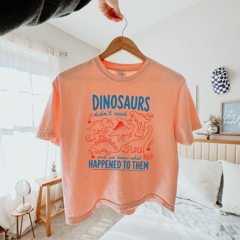 May include: A peach-colored t-shirt with the text "DINOSAURS didn't read and we know what HAPPENED TO THEM" in blue. The shirt features a red illustration of various dinosaurs and books. The shirt is hanging on a wooden hanger.