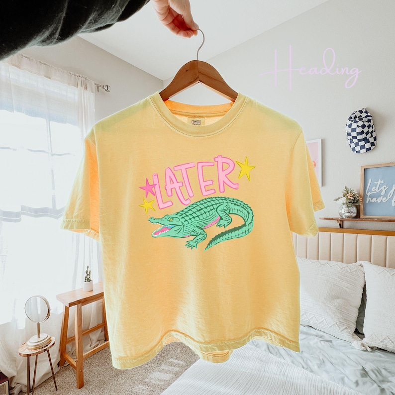 Peut inclure: T-shirt jaune avec le mot "LATER" en rose et un dessin d'alligator vert. Le t-shirt est suspendu &agrave; un cintre en bois. L'alligator a des d&eacute;tails roses et des &eacute;toiles jaunes.