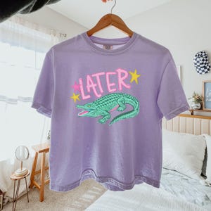 Peut inclure: Un t-shirt lavande avec un motif d'alligator vert et le mot "LATER" en rose avec des &eacute;toiles jaunes. Le t-shirt &agrave; manches courtes est suspendu &agrave; un cintre en bois.