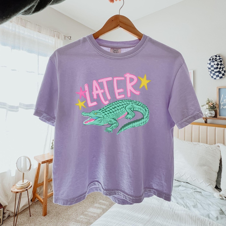 Peut inclure: Un t-shirt lavande avec un motif d'alligator vert et le mot "LATER" en rose avec des &eacute;toiles jaunes. Le t-shirt &agrave; manches courtes est suspendu &agrave; un cintre en bois.