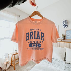 May include: A peach-colored t-shirt with the text "PROPERTY OF BRIAR UNIVERSITY -XXL- ATHLETIC DEPARTMENT" in navy blue. The shirt is hanging on a wooden hanger.