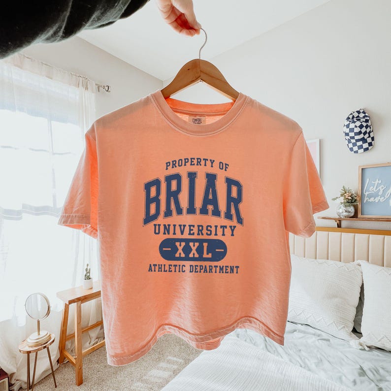 May include: A peach-colored t-shirt with the text "PROPERTY OF BRIAR UNIVERSITY -XXL- ATHLETIC DEPARTMENT" in navy blue. The shirt is hanging on a wooden hanger.