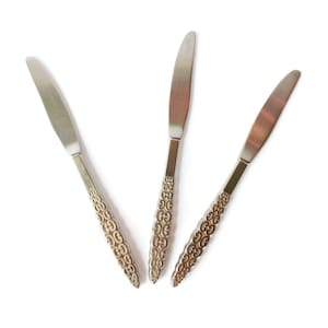 Peut inclure: Trois couteaux à beurre en argent avec des manches ornés. Les couteaux sont disposés en forme de V.