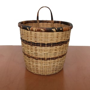 Op de afbeelding: Een geweven rieten mand met een bruin en zwart gestreept patroon en een handvat. De mand staat op een houten oppervlak.