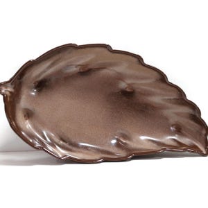 Puede incluir: Una fuente de cerámica con forma de hoja. El plato presenta un degradado de tonos marrones, con un borde marrón más oscuro y un centro más claro. El diseño de la hoja tiene detalles texturizados y un acabado brillante.