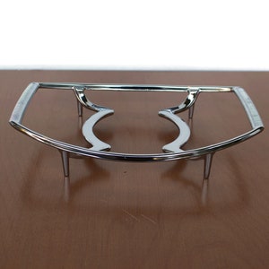 Könnte beinhalten: Ein verchromter Metall-Topfuntersetzer mit gebogenem Design und drei Beinen. Der Untersetzer ist so konzipiert, dass er Geschirr erhöht und Oberflächen vor Hitze schützt.