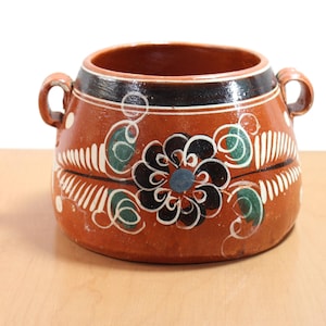 Puede incluir: Una olla de cerámica de color terracota con dos asas. La olla tiene un borde negro y está decorada con una gran flor negra con un centro azul, detalles blancos y espirales verdes. La olla se encuentra sobre una superficie marrón claro.